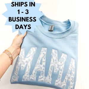 May include: Light blue sweatshirt with the word "MAMA" in white with a floral pattern. The sweatshirt is folded and held by a hand.