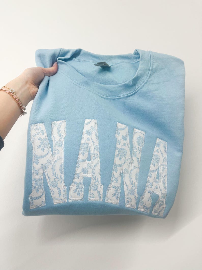 May include: Light blue sweatshirt with the word "NANA" in white with a floral pattern.