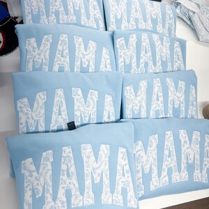 May include: Six light blue sweatshirts with the word "MAMA" printed in white with a white floral pattern. The word "MAMA" is in a large, bold font.