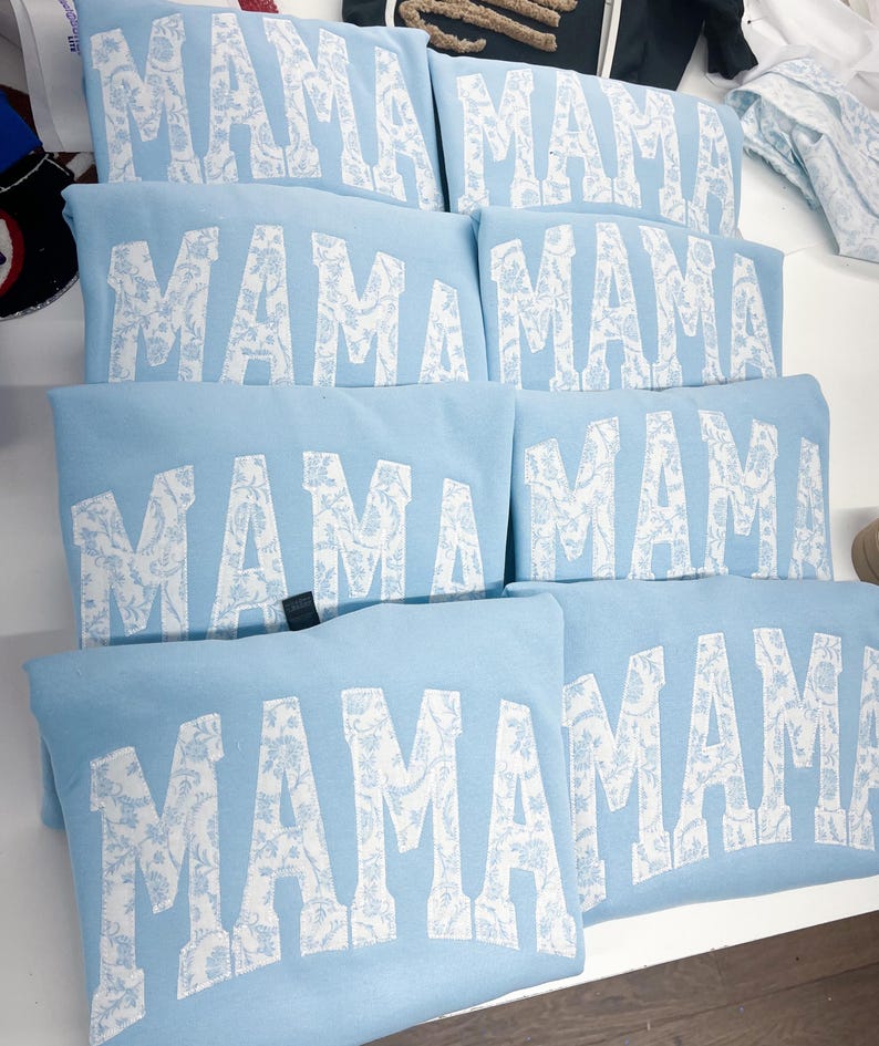 May include: Six light blue sweatshirts with the word "MAMA" printed in white with a white floral pattern. The word "MAMA" is in a large, bold font.