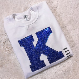 May include: White t-shirt with a large blue sequined letter K on the front. The shirt is folded and has a size tag that says "Medium" on it.