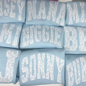 May include: A stack of light blue sweatshirts with white lace-like lettering. The names on the sweatshirts are Chase, Nana, Mimi, Kim, Biggie, Bes, J, Sonny, and Bubble.