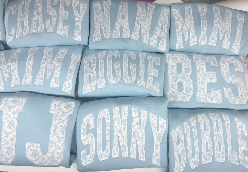 May include: A stack of light blue sweatshirts with white lace-like lettering. The names on the sweatshirts are Chase, Nana, Mimi, Kim, Biggie, Bes, J, Sonny, and Bubble.