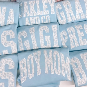 May include: A stack of light blue sweatshirts with white patterned lettering. The lettering includes the words "NANA", "RES", "GIGI", "PRES", "OAKLEY AND CO", "GRAY", "BOY MOM", and "BEBE".