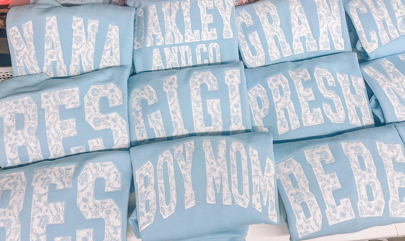 May include: A stack of light blue sweatshirts with white patterned lettering. The lettering includes the words "NANA", "RES", "GIGI", "PRES", "OAKLEY AND CO", "GRAY", "BOY MOM", and "BEBE".