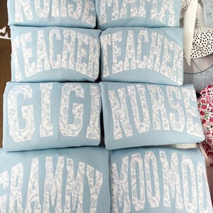 May include: Six light blue sweatshirts with white text and a floral pattern. The text on the sweatshirts reads "Nurse", "Mama", "Teacher", "Teacher", "Gigi", "Nurse", "Grammy", and "MooMoo".