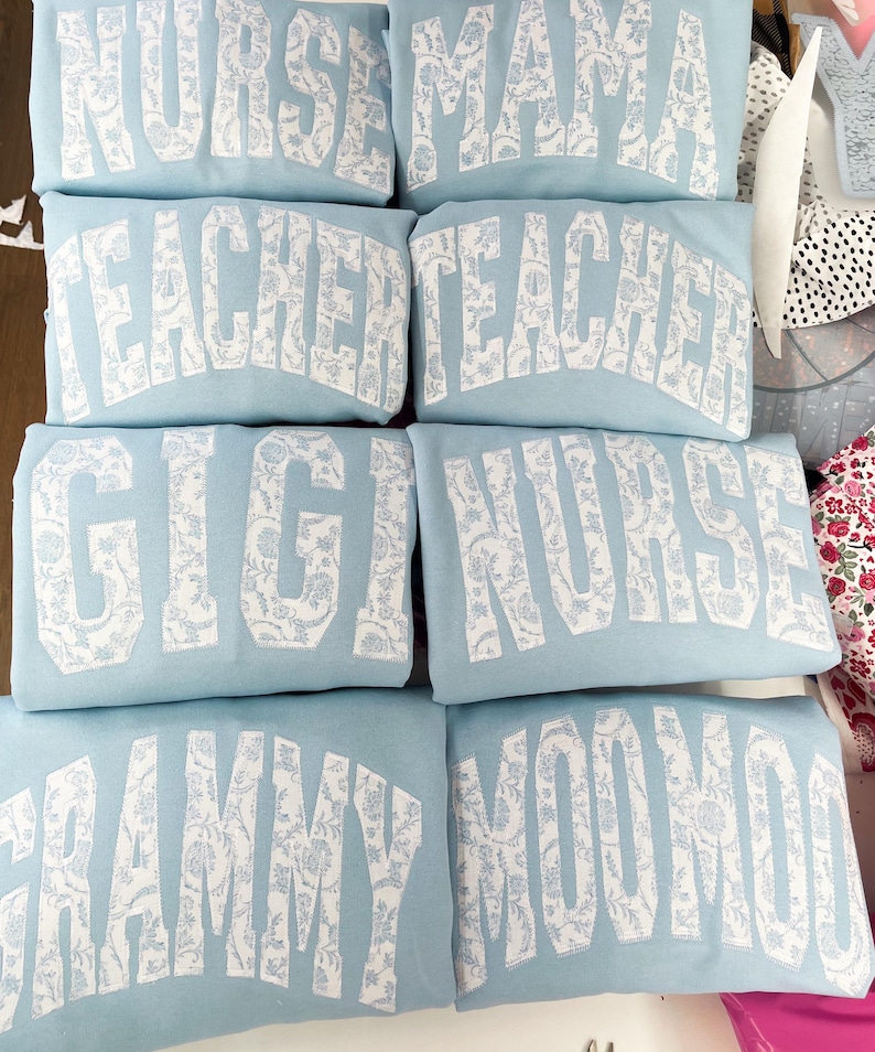 May include: Six light blue sweatshirts with white text and a floral pattern. The text on the sweatshirts reads "Nurse", "Mama", "Teacher", "Teacher", "Gigi", "Nurse", "Grammy", and "MooMoo".