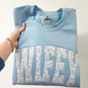 May include: A light blue sweatshirt with the word "WIFEY" in white with a floral pattern on the letters.