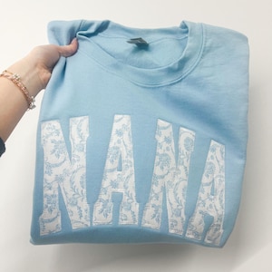 May include: Light blue sweatshirt with the word "NANA" in white with a floral pattern.