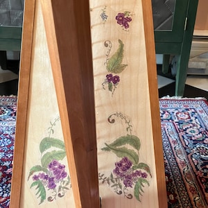 May include: A wooden harp with a light wood finish and floral designs painted on the soundboard. The harp has a natural wood color and is decorated with purple and green flowers.
