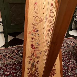 Pode incluir: Uma harpa de madeira com uma caixa de ressonância de cor clara e uma moldura de madeira escura. A caixa de ressonância é decorada com um desenho floral em tons de vermelho, laranja e castanho. As cordas da harpa são visíveis.