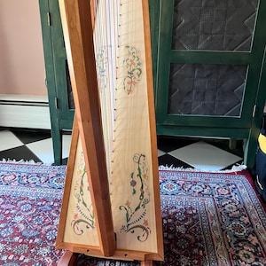 May include: A wooden harp with a light-coloured soundboard and a dark wood frame. The soundboard is decorated with floral designs in green, yellow, and orange. The harp has multiple strings in various colours and is set on a wooden stand.
