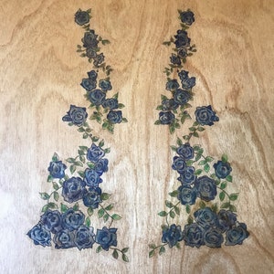 Op de afbeelding: Twee symmetrische ontwerpen van blauwe rozen en groene bladeren op een licht houten oppervlak.