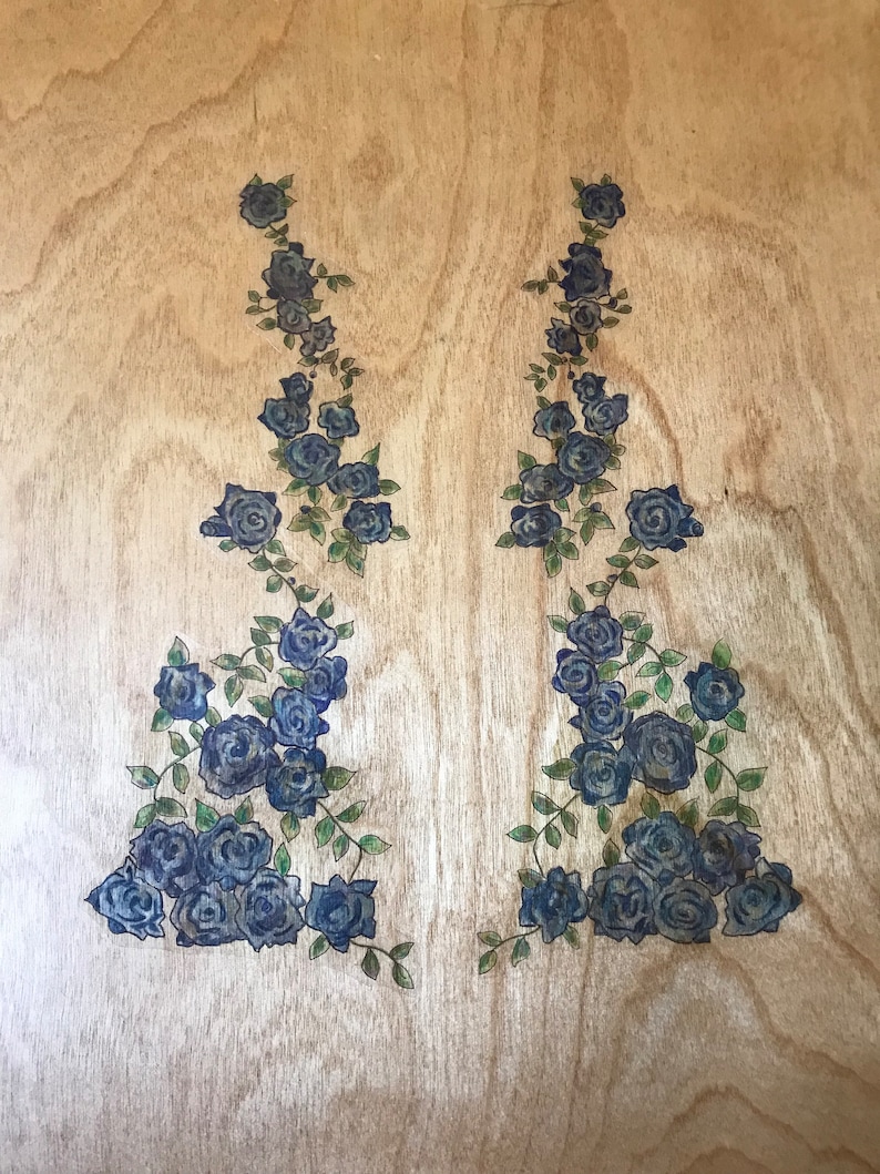 Op de afbeelding: Twee symmetrische ontwerpen van blauwe rozen en groene bladeren op een licht houten oppervlak.