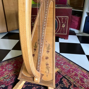 May include: A wooden harp with a natural finish and a floral design on the soundboard. The harp has a stand with three legs.