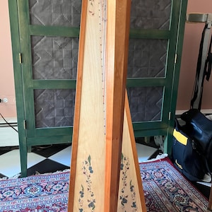 Puede incluir: Un arpa de madera con una tabla armónica de madera clara y un marco de madera oscura. La tabla armónica está decorada con diseños florales en azul y verde. El arpa tiene cuerdas de varios colores y está sobre una alfombra estampada.