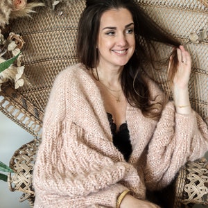 May include: A light pink, open-front cardigan with textured sleeves. The cardigan is made of a soft, fuzzy material. The woman is sitting in a wicker chair. She is wearing a black top and black pants. She is also wearing gold jewelry.