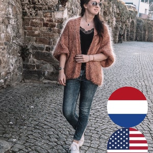 Puede incluir: Un cárdigan abierto y difuso de color melocotón con mangas anchas, usado sobre una camiseta negra con ribete de encaje y jeans azul oscuro. El conjunto se completa con zapatillas blancas, un collar plateado con un colgante oscuro y gafas de sol plateadas.