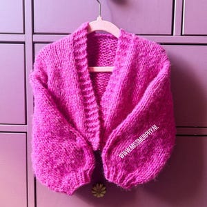 May include: A bright pink chunky knit cardigan with long sleeves and a ribbed collar. The garment is displayed on a light pink hanger. The text WWW.MISSMURPHY.NL is printed in white on the lower right sleeve of the knitted fabric.