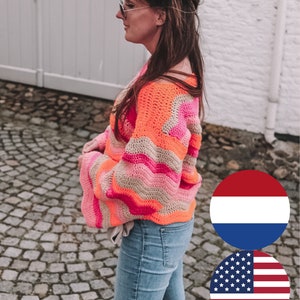 Op de afbeelding: Een kleurrijke gehaakte trui in oranje, roze en beige. De trui heeft een golvend patroon en lange mouwen. De persoon draagt een blauwe spijkerbroek en een zonnebril.