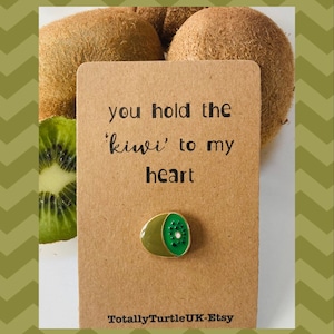 May include: A brown card with the text "you hold the 'kiwi' to my heart" and a gold pin with a green kiwi fruit design. The card is surrounded by kiwi fruit.