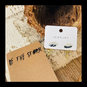 Peut inclure: Une paire de boucles d'oreilles en or avec des breloques en forme de nuage noir et blanc et des accents de foudre et de cristal pendants. Les boucles d'oreilles sont présentées sur une carte blanche avec le mot "JEWELLRY" imprimé dessus. La carte est posée sur une surface en bois. Le texte "BE THE STORM" est imprimé sur une carte brune.