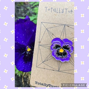 Puede incluir: Un broche de flor de pensamiento morado y negro sobre una tarjeta marrón con el texto "Totally Tuk". El broche tiene un contorno dorado y un centro amarillo. Una flor de pensamiento morada real en el fondo.