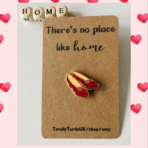 Peut inclure: Une carte brune avec le texte "There's no place like home" et une paire de chaussons de rubis rouges à paillettes. La carte comporte également le texte "HOME" en lettres de bois et le texte "Totally TurtleUK/shop/etsy".