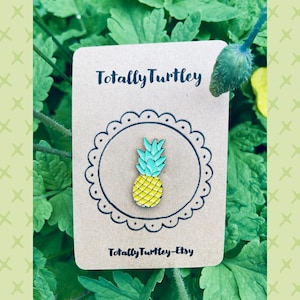 Op de afbeelding: Een kleurrijke emaille pin met een geel en turquoise ananasontwerp. De pin zit op een bruine kaart met de tekst "Totally Turtley" en "Totally Turtley-Etsy". De achtergrond is groen gebladerte.