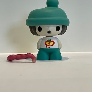 May include: A small, teal blue figurine of a person wearing a teal blue hat with a white pom-pom. The figurine has a white shirt with a red apple graphic and teal blue pants. A pink worm is lying on the white surface.