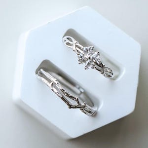 以下が含まれることがあります： 白い六角形のスタンドに展示された2つのシルバーリング。1つのリングは透明な宝石が付いた花の模様で、もう1つは枝のような質感です。リングは清潔で明るい背景に配置されています。