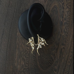 May include: Gold-coloured earrings with a wing-like design, set with small, clear gemstones. The earrings are displayed on a black ear-shaped form against a dark wood-grain background.