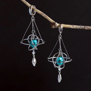 May include: A pair of silver-toned earrings with a celestial design. Each earring features a geometric frame with a teal-colored orb at the center, accented by dangling crystal and diamond-shaped charms. The earrings are suspended from a thin branch against a dark background.