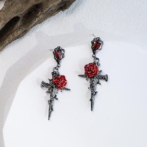 May include: A pair of gothic earrings. Each earring has a black cross design with a red rose at the centre and a red gemstone at the top. The earrings have a dark, antique finish, creating a striking contrast.