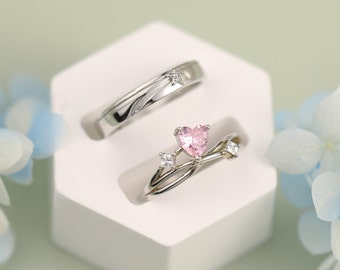 May include: A pair of silver rings with a pink heart-shaped stone and small clear stones on one ring and a star-shaped stone on the other ring. The rings are displayed on a white hexagonal ring holder.