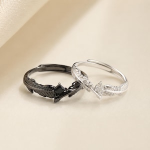May include: Two adjustable rings featuring a feather design. One ring is finished in black with a central black square-cut stone. The other ring is finished in silver with a central clear marquise-cut stone and small clear stones along the band.