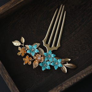 May include: A gold-coloured hair comb with a floral design. The comb has a series of tines and is decorated with turquoise and orange flowers, and gold-coloured leaves. It is set against a dark wooden background.