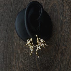 Puede incluir: Pendientes de oreja dorados con un diseño delicado en forma de ala. Los pendientes presentan una serie de pequeñas piedras preciosas transparentes engastadas a lo largo de las líneas curvas de las alas. Se exhiben sobre un fondo de madera oscura.