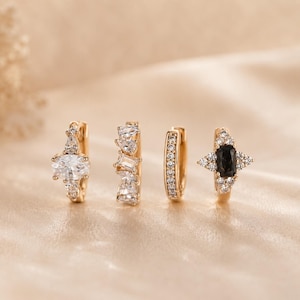 Könnte beinhalten: Ein Set aus vier goldenen Creolen. Die erste hat einen ovalen klaren Stein, die zweite eine Ansammlung von klaren Baguette- und Rundsteinen, die dritte ist eine einfache Pavé-Creole und die vierte zeigt einen rechteckigen schwarzen Stein mit Akzenten.