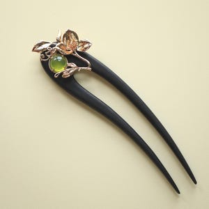 May include: A black wooden hair fork with a decorative gold-colored floral design. The floral design includes a green gemstone and gold-colored leaves. The hair accessory is a two-pronged fork.