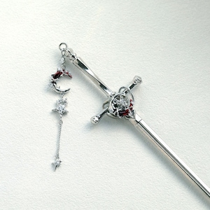 Pode incluir: Um acessório de cabelo prateado e um pingente pendurado. O acessório de cabelo apresenta um design de espada com uma gema vermelha e um detalhe de estrela. O pingente inclui uma lua crescente, uma estrela e estrelas penduradas, todas com gemas claras.