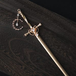 May include: A gold-colored hair stick with a sword-like design. The stick features a decorative cross-shaped element with a square gemstone and a dangling crescent moon charm with clear crystals. The hair accessory is set against a dark wood surface.