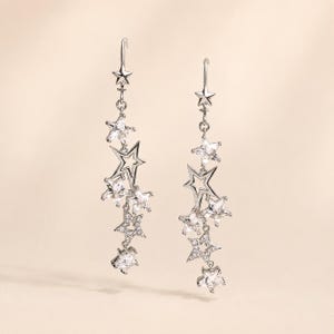 May include: A pair of silver-toned drop earrings featuring a cascading design of multiple star-shaped charms. Each earring includes a mix of solid metal stars, open-frame stars, and stars encrusted with clear, sparkling stones suspended from a hook.