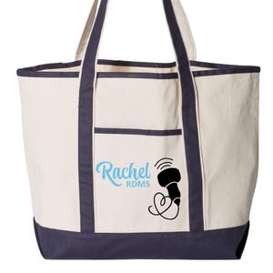 Peut inclure: Sac fourre-tout crème avec garniture et poignées bleu marine. Le sac présente une poche avant et un motif avec le nom "Rachel" en écriture bleu clair, ainsi qu'un dessin noir d'un appareil médical.