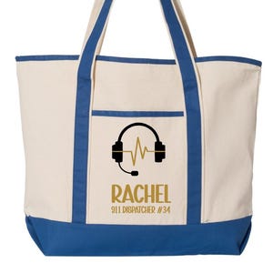Könnte beinhalten: Cremefarbene Tragetasche mit blauen Akzenten. Mit einer schwarzen Grafik von Kopfhörern und einer Schallwelle, mit dem Namen "RACHEL" und "911 DISPATCHER #34" in goldfarbenen Buchstaben. Die Tasche hat blaue Griffe und einen blauen Boden.