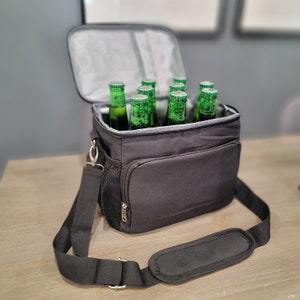 May include: A black insulated cooler bag with a zippered pocket and adjustable shoulder strap. The bag is filled with green glass bottles.