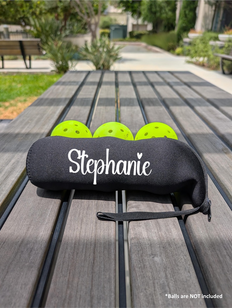 Peut inclure: Une housse de raquette de pickleball en n&eacute;opr&egrave;ne noir avec le nom "Stephanie" en &eacute;criture blanche. Trois balles de pickleball vert fluo sont visibles. La housse a une sangle et une fermeture &eacute;clair noires. Le fond est une table en bois dans un parc.