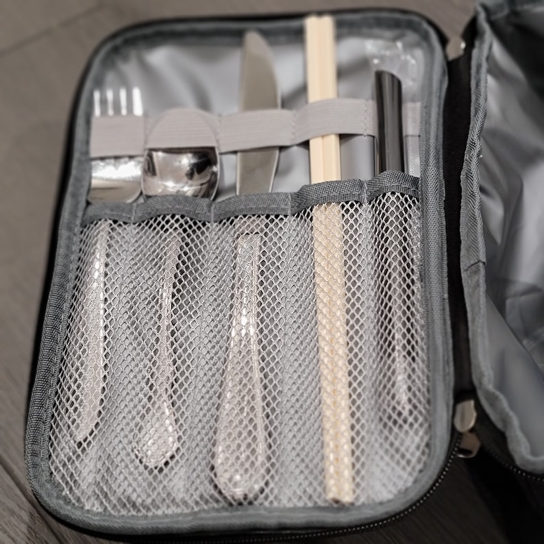 Puede incluir: Una maleta de viaje negra con cierre de cremallera, que contiene un juego de cubiertos de plata y palillos. Los cubiertos se guardan en una bolsa de malla con ranuras individuales para cada pieza.