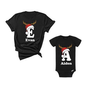 May include: Black t-shirts and a baby onesie with Christmas designs. The t-shirt has a white letter "E" with a reindeer antler design and a Santa hat, with the name "Evan" below. The onesie has a white letter "A" with the same design and the name "Aiden".