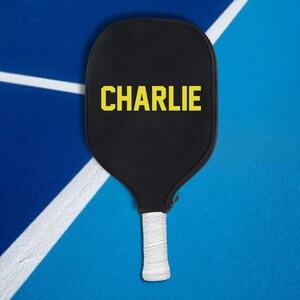 May include: A black pickleball paddle with a white grip and the name "CHARLIE" printed in yellow letters.